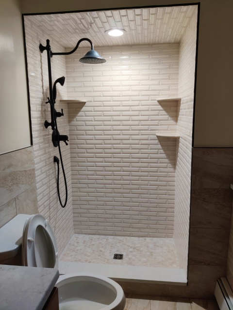 Sophisticated bathroom renovation completed by True Built Construction showcasing an upscale walk-in shower with textured beige herringbone tiles, sleek black shower fixtures, and dual inset niches for amenities. The design reflects a modern aesthetic with a nod to classic elegance