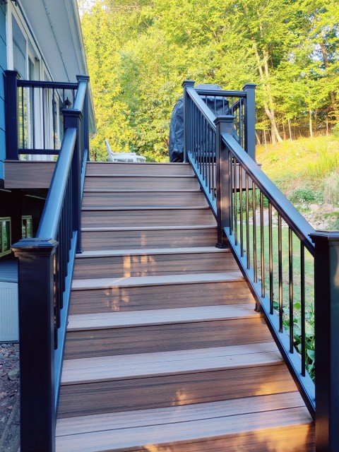 Custom-designed outdoor deck built by True Built Construction featuring rich walnut-toned planks with a smooth finish, paired with contrasting deep blue railing and balusters. Elegant stairs descend to a verdant, tree-lined backyard, highlighting quality craftsmanship and design