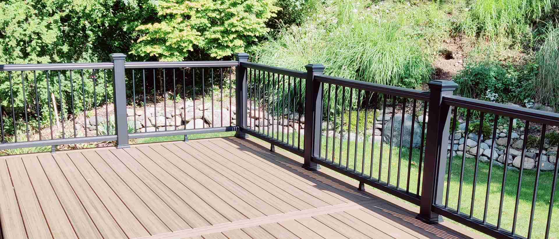 A full deck rebuild by True Built Construction features a spacious, multi-tier design with luxurious walnut-colored composite planks and contrasting black aluminum balusters, all set against the serene backdrop of a dense, leafy forest