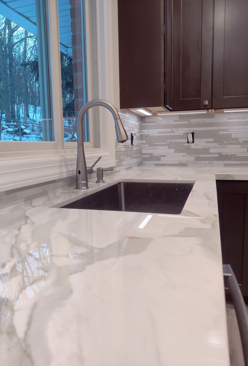 Elegant kitchen sink area remodeled by True Built Construction, showcasing a deep, undermount sink with a high-arc faucet, set against a luxurious marbled white countertop. The linear mosaic tile backsplash in shades of grey adds sophistication, while the window frames a serene, snowy landscape